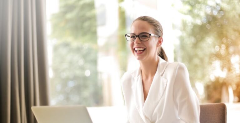 Cheerful businesswoman in glasses working on a laptop, in a bright and modern office setting.